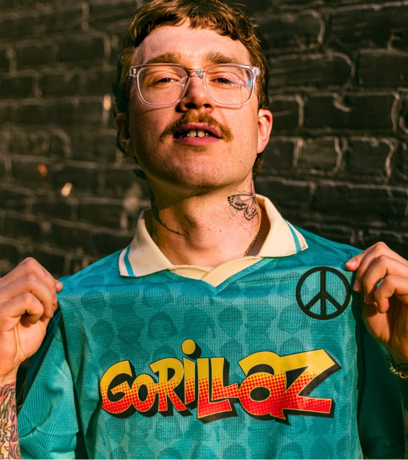 A man wearing a teal Gorillaz band t-shirt with a peace sign and the band's name in yellow and red, posing in front of a dark brick wall.
