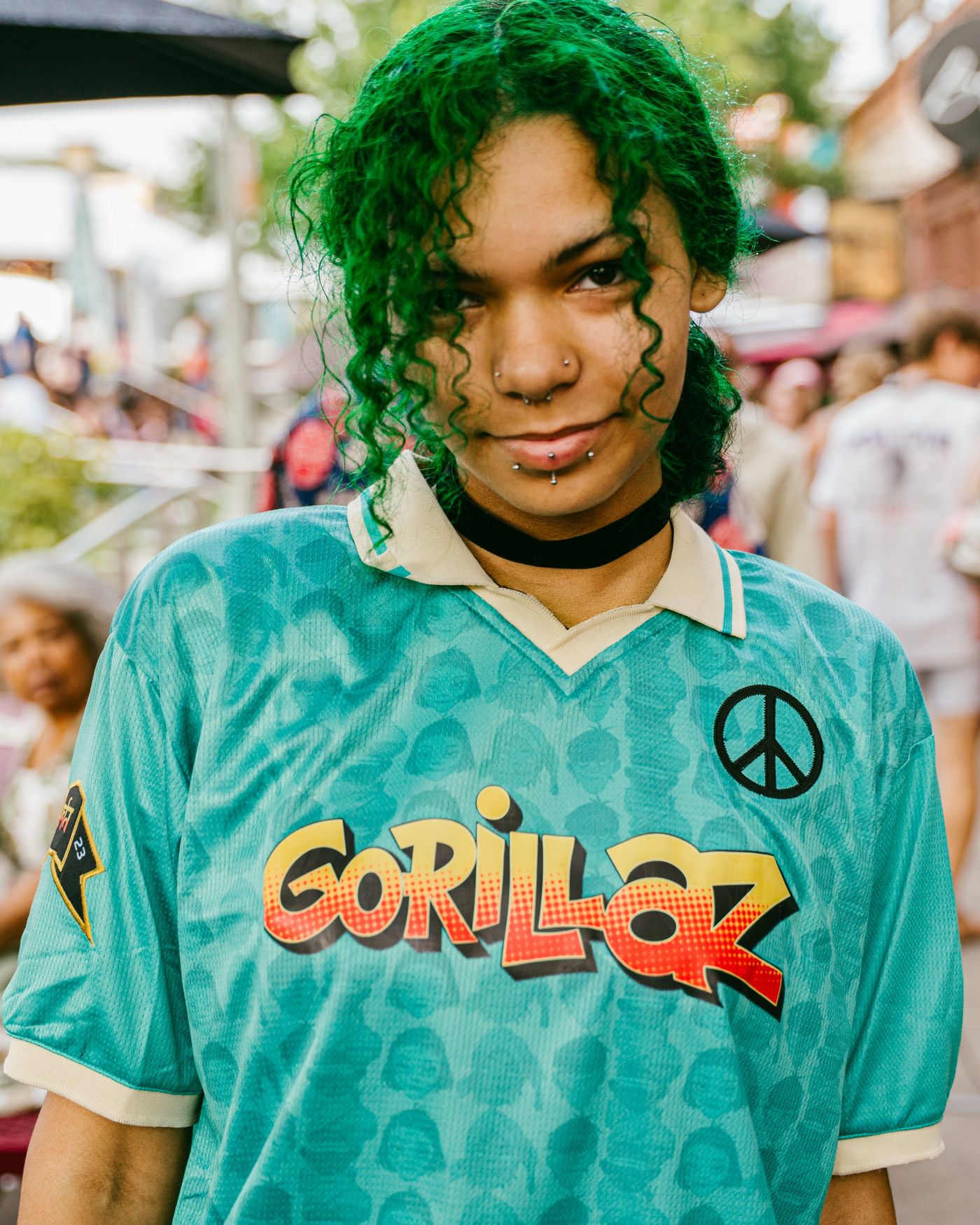A person outdoors wearing a turquoise Gorillaz band t-shirt with a camouflage pattern, featuring the band's name in red and yellow gradient letters and a black peace sign on the chest.